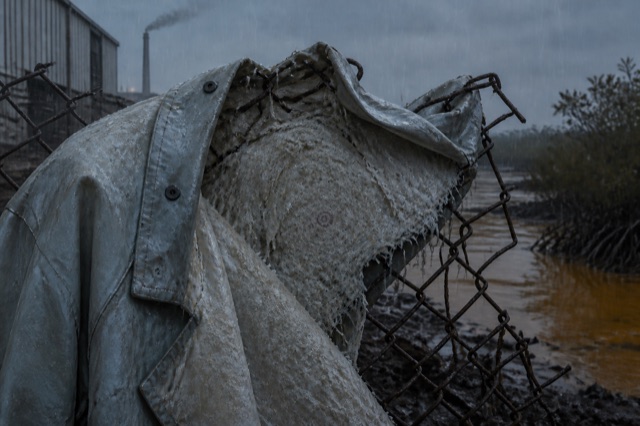 A pale biofabric coat hangs across a broken rusted fence in heavy rain, its raw inner lining marked by a small bruise-like rosette between a textile factory and muddy mangroves.
